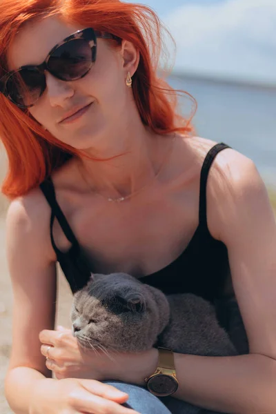 Red Hair lady holds beautiful Grey Scottish-fold shorthair fluffy cat with orange eyes outside near the river. Warm picture toning. Pets care. World domestic animal day. Image for websites about cats.