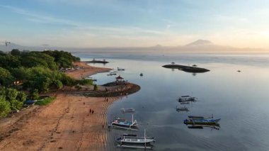 Güneşin doğuşunda arka planda renkli balıkçı tekneleri, çardaklar ve Agung Dağı 'nın olduğu huzurlu sahilin insansız hava aracı görüntüsü. Tropikal dinginliği ve Bali sahil cazibesini yakalar.