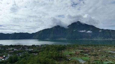 Endonezya, Bali 'deki Batur Dağı ve Batur Gölü' nün insansız hava aracı görüntüleri. Etrafı yeşil orman, tarım arazisi ve bulutlu gökyüzünün altındaki dağınık köylerle çevrili. Çarpıcı bir volkanik manzara..