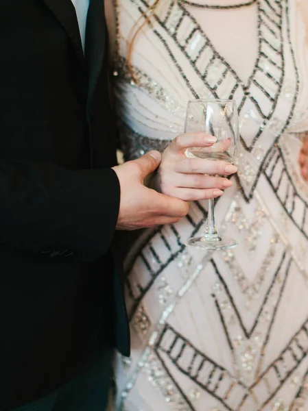 平和的なビーズのパターンを持つアートデコのドレスの花嫁は白ワインのボサールを保持しています 黒いスーツの新郎は花嫁の手を握っている 結婚式日 — ストック写真