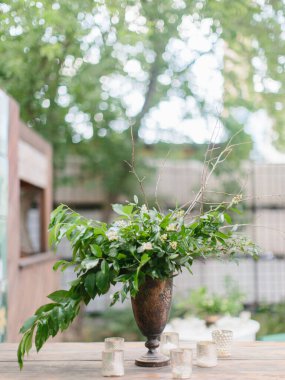 Bir buket yeşil yaprak, beyaz çiçekler ve dallar antika bir bakır vazonun içinde duruyor. Cam şamdanlar masanın üzerinde. Açık hava parti dekorasyonu.