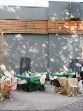 Yeşil renkte düğün ziyafeti ve açık havada altın rengi. Çiçek buketleri, masalarda renkli sandalyeler. Arka planda ahşap dekorlu beton bir duvar var..