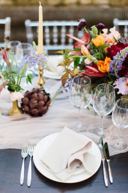 Mavi-beyaz düğün ziyafeti masası. Ön planda peçeteli, şarap kadehli, bıçaklı ve çatallı beyaz tabaklar var. Parlak çiçeklerin arkasında. Tüllü kumaş..