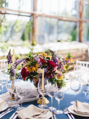 Serada düğün ziyafeti masası servisi. Mavi bir masa örtüsü, parlak kır çiçekleri, şarap bardakları, beyaz tabaklar, çatal bıçaklar. Panoramik pencerelerin arkasında.
