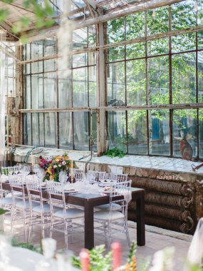 Serada ziyafet masası servis ettim. Çiçekli buketler, bir bardak şarap, tabaklar masanın üstünde. Arka planda bir panorama penceresi ve park manzarası var..