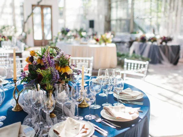 Serving a wedding banquet table in the greenhouse. On a blue tablecloth, a bouquet of bright wild flowers, wine glasses, white plates, cutlery, candles in golden candlesticks.