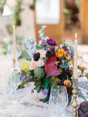 Tropik bir anthurium buketi, enginar, ziyafet masasında laleler. Ön planda şarap kadehleri, altın şamdanlarda yanan mumlar var..