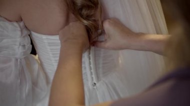 Gelinliğin çıkarılması, gelinliğin ayrıntıları. Gelin bir gelinlik giyiyor. Annem korsenin bağlanmasına yardım ediyor. Gelinliğe kurdele bağlayan nedime. Yavaş çekim.