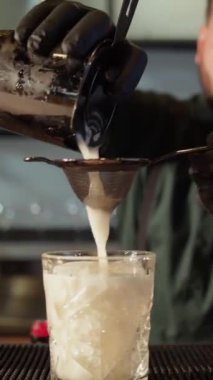 Profesyonel bir barmen alkollü kokteyl hazırlar. Bir barmen bardaktan eleğe kokteyl döker. Kokteyl beyaz. Dikey video.