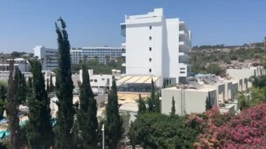 Ayia Napa, Kıbrıs - 6 Haziran 2023: Kıbrıs Rum Kesimi 'nin Ayia Napa kentindeki bir otelden manzara, beyaz binalar, çiçek açan bahçeler, yeşil palmiye ağaçları ve parlak mavi bir gökyüzünün altında manzaralı bir tepe.