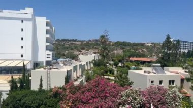 Ayia Napa, Kıbrıs - 6 Haziran 2023: Kıbrıs Rum Kesimi 'nin Ayia Napa kentindeki bir otelden manzara, beyaz binalar, çiçek açan bahçeler, yeşil palmiye ağaçları ve parlak mavi bir gökyüzünün altında manzaralı bir tepe.