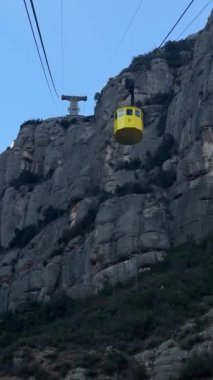 Montserrat, İspanya - 6 Aralık 2023: Santa Maria de Montserrat Manastırı, Monistrol de Montserrat, Katalonya. Sarı teleferik mavi gökyüzünde yüksek bir dağa doğru gidiyor..