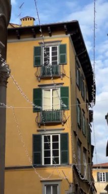 Lucca, İtalya - 27 Kasım 2024: Lucca 'daki geleneksel İtalyan binası, sarı duvarlar, yeşil kepenkler ve işlenmiş demir balkonlarla süslenmiş, açık mavi gökyüzü altındaki şenlik iplikleriyle süslenmiş.