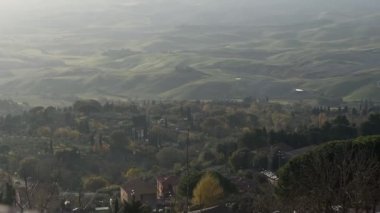 Toskana kırsalının yuvarlanan yeşil tepeleri Volterra 'dan görülebiliyor. Kırsal binalar ve ön planda uzun selvi ağaçları yumuşak sisli bir gökyüzünün altında..