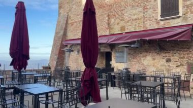 Montepulciano, İtalya - 25 Kasım 2024: Montepulciano 'da tuğla duvarlar, kapalı bordo şemsiyeler, siyah metal mobilyalar ve parlak gökyüzü altında panoramik kırsal manzaralı açık kafe terası.