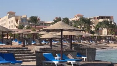 Hurghada, Mısır - 11 Kasım 2024: Hurghada 'da bir tatil köyü manzarası. Hasır sıralar, mavi güneş şemsiyeleri ve palmiye ağaçları rahatlatıcı bir atmosfer yaratıyor. Kızıl Deniz kıyısında sakin bir tatil..