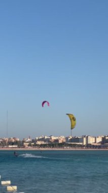 Hurghada, Mısır - 5 Kasım 2024: Uçurtma sürücüsü turkuvaz denizi boyunca süzülerek açık mavi gökyüzünün altında bir kıyı şehri yakınlarında su sıçratır.