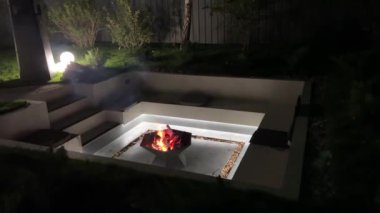 Geceleri yeşilliklerle çevrili geometrik tasarımı olan modern bahçe ateşi çukuru. Sıcak ışık ve minimalist dekorla rahat ve şık bir açık hava rahatlama alanı..