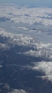 Bir uçak penceresinden görülen, karla kaplı Alp dağlarının çarpıcı manzarası. Yüksek tepeler bulutların üzerinde yükselir, huzurlu ve nefes kesici bir kış manzarası yaratır..
