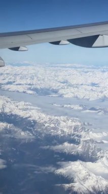Karla kaplı dağ sırasının üstündeki kanadı gösteren uçak penceresinden bak. Açık mavi gökyüzü ve çarpıcı kış manzarası uçuş sırasında yüksek irtifadan.