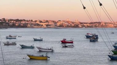 Portekiz 'in Cascais şehrinde günbatımında sakin sularda yüzen balıkçı tekneleri. Şehrin ufuk çizgisi arka planda görülebilir, sıcak altın ışıkla yıkanmış. Renkli tekneleri olan huzurlu kıyı manzarası.