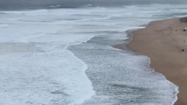 Büyük okyanus dalgaları Portekiz 'deki Praia do Norte kumsalına vuruyor. Okyanus güçlü ve vahşi görünüyor, ve kıyı gri gökyüzünün altında uzanıyor..