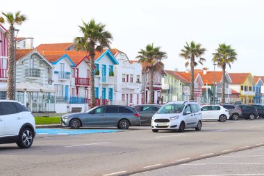Aveiro, Portekiz - 20 Şubat 2025: Kosta Nova 'da palmiye ağaçları ve cadde boyunca park etmiş arabalarla renkli çizgili evler, canlı mimari ve sahil cazibesiyle kıyı manzarası yaratıyor.
