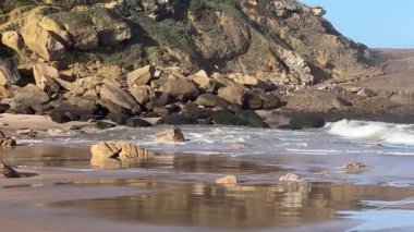 Okyanus dalgalarının yavaşça kıyıya vurduğu güzel kumlu bir sahil. Yeşil bitki örtüsüyle kaplı büyük kayalar ve kayalıklar çarpıcı bir doğal zemin oluşturur. Huzurlu ve manzaralı bir kıyı manzarası.