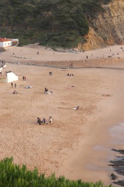 İnsanlar Portekiz 'deki kumlu Praia do Magoito sahilinde huzurlu bir günün tadını çıkarıyorlar. Etrafı kayalık kayalıklarla ve yeşil tepelerle çevrili. Dalgalar yavaşça kıyıya ulaşıyor..