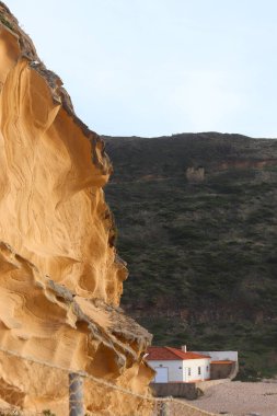 Praia do Magoito, Portekiz 'de günbatımı ışıklarıyla aydınlatılan altın kumtaşı uçurumu, sahil yakınlarındaki engebeli yeşil tepelerin altına kurulmuş beyaz bir ev ve kırmızı bir çatı..