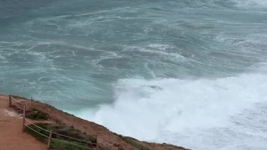 Güçlü okyanus dalgaları Nazare, Portekiz 'in kayalıklarına çarpar. Dünyanın en büyük dalgaları bulutlu bir kış gününde manzaralı bir uçurumdan yakalanır..