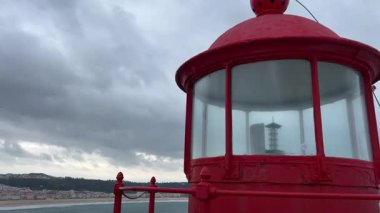 A red lighthouse at Forte de Sao Miguel Arcanjo in Nazare, Portugal. The historic beacon stands against a cloudy sky, overlooking the Atlantic Ocean, guiding sailors near the famous big wave surf spot