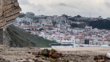 Portekiz, Nazare 'nin deniz feneri bölgesinden görünüşü. Beyaz evleri ve kumsalda kırmızı çatıları olan bulutlu gökyüzünün altında bir sahil kasabası..