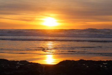 Portekiz, Praia do Magoito üzerinde çarpıcı bir günbatımı. Altın güneş okyanus dalgalarına yansıyarak barışçıl ve sıcak bir atmosfer yaratıyor. Gökyüzü turuncu renklerle dolu, dalgalı denizle harmanlanmış..