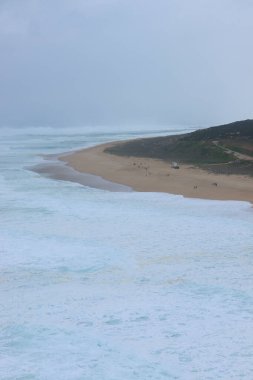 Büyük okyanus dalgaları Portekiz 'deki Praia do Norte kumsalına vuruyor. Okyanus güçlü ve vahşi görünüyor, ve kıyı gri gökyüzünün altında uzanıyor..