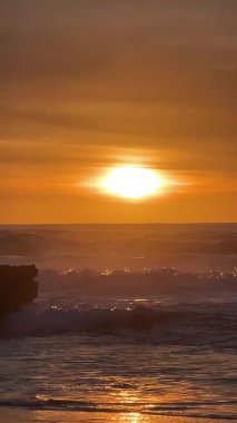 Portekiz, Praia do Magoito üzerinde çarpıcı bir günbatımı. Altın güneş okyanus dalgalarına yansıyarak barışçıl ve sıcak bir atmosfer yaratıyor. Gökyüzü turuncu renklerle dolu, dalgalı denizle harmanlanmış..
