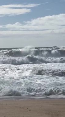 Güçlü okyanus dalgaları, Portekiz, Nazar 'da parçalı bulutlu bir gökyüzünün altında kumlu sahile çarpıyor ve Atlantik kıyısının dinamik güzelliğini gösteriyor..