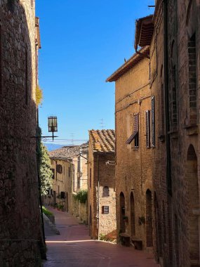 İtalya, San Gimignano 'da dar bir ortaçağ caddesi. Taş binalar, mavi gökyüzü ve sıcak güneş ışığı huzurlu ve tarihi bir atmosfer yaratıyor..