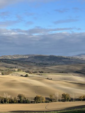 Pienza, İtalya - 25 Kasım 2024: Tuscan kırsalının hareketli tepeleri, sonbahar yaprakları, bulutlu gökyüzü altındaki uzak dağlar. İtalya 'nın huzurlu kırsal güzelliğinin mükemmel bir temsili.
