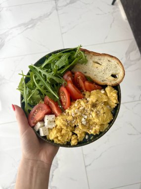Modern bir mutfakta, çırpılmış yumurta, ekmek tostu, domates ve roka yeşil bir tabakta bir kadının elinde. Sağlıklı kahvaltı kavramı.