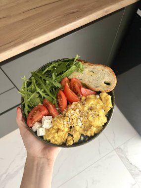 Modern bir mutfakta, çırpılmış yumurta, ekmek tostu, domates ve roka yeşil bir tabakta bir kadının elinde. Sağlıklı kahvaltı kavramı.