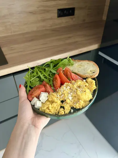 Modern bir mutfakta, çırpılmış yumurta, ekmek tostu, domates ve roka yeşil bir tabakta bir kadının elinde. Sağlıklı kahvaltı kavramı.