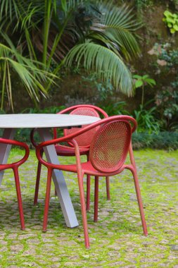 Kafe tarzı bir bahçede, arka planda palmiye ağaçları ve yemyeşil yemyeşil bir masa etrafında kırmızı bir set..