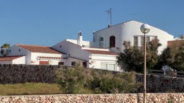 Beyaz Akdeniz tarzı çatı kiremitli bir evin manzarası. Cap d Artrutx, Menorca 'da açık mavi bir gökyüzüne karşı kurulmuş. Taş duvar ve yeşil bitkiler dingin atmosfere eklenir.