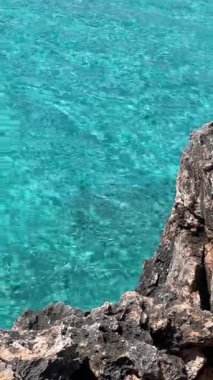 Berrak turkuaz su, güneş ışığı altında parıldıyor, kıyıda engebeli karanlık kayalar var..