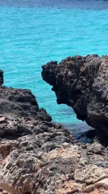 Berrak turkuaz su, güneş ışığı altında parıldıyor, kıyıda engebeli karanlık kayalar var..