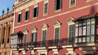 Cuitadella de Menorca 'nın büyüleyici mimarisinin tarihi bir görüntüsü. Altın güneş ışığı altında balkonları ve kemerleri olan klasik kırmızı binalar sergileniyor..