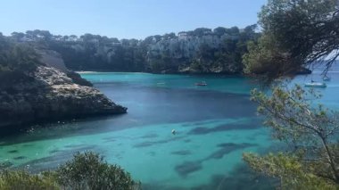 İspanya 'nın Cala Galdana kentinin berrak turkuaz suları ve sakin denizde yavaşça yüzen tekneleriyle nefes kesici manzarası. Çevredeki kayalıklar ve yeşillikler körfezin doğal güzelliğini arttırır..