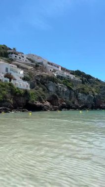 Cala en Porter, Menorca 'daki kristal gibi sığ sular, parlak mavi gökyüzünün altında kayalık bir uçuruma tünemiş beyaz evler. Güneşli bir günde huzurlu bir sahil köyü.