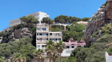 Cala en Porter, Menorca 'daki parlak mavi gökyüzüne bakan yemyeşil ve palmiye ağaçlarıyla çevrili bir tepeye tünemiş beyaz ve pembe evlerin manzarası..